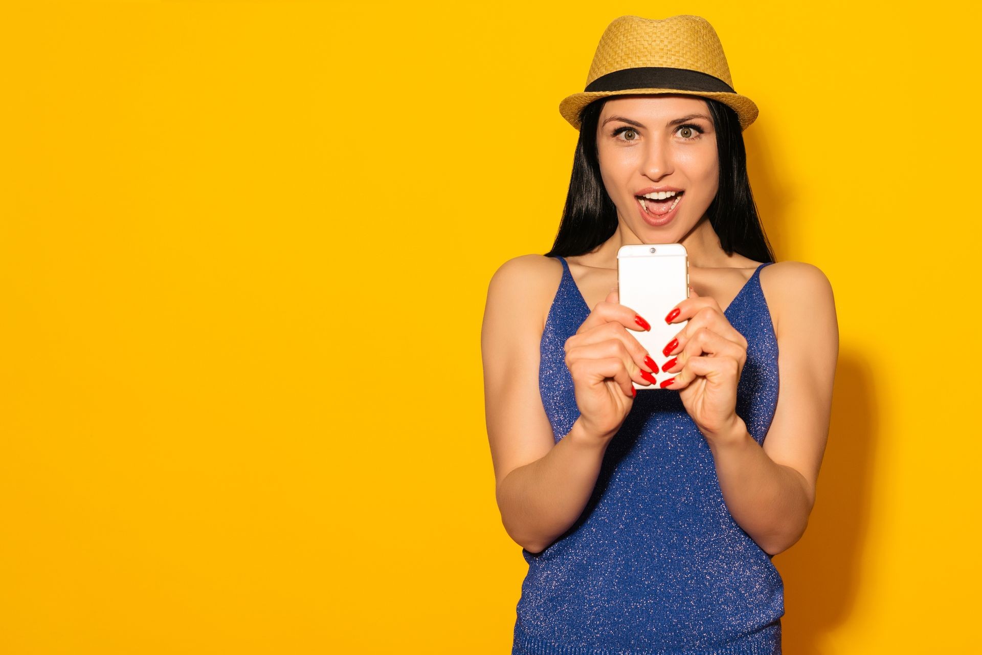 Excited laughing woman in hat standing and using mobile phone over yellow background - Image